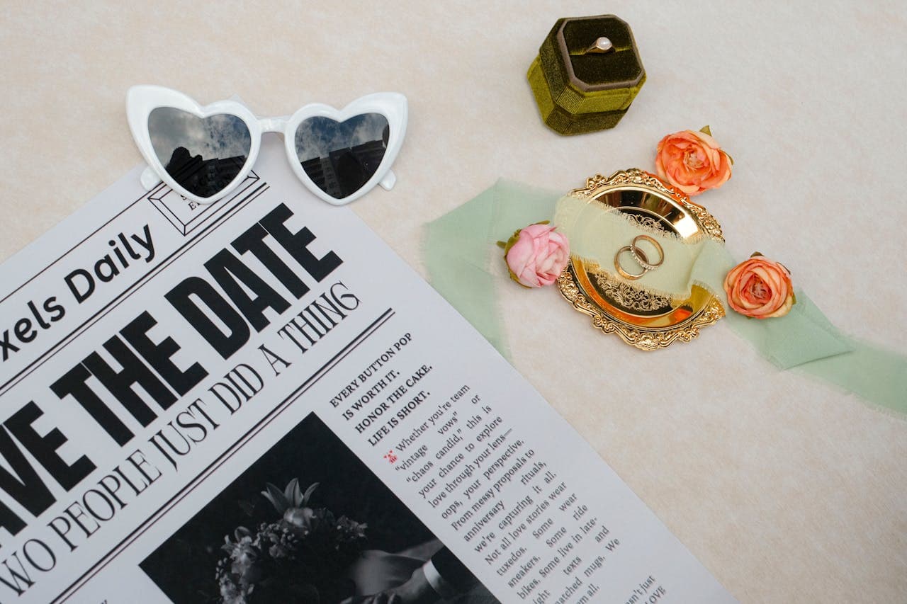 Flat lay with a newspaper-style wedding announcement, rings, and heart-shaped sunglasses