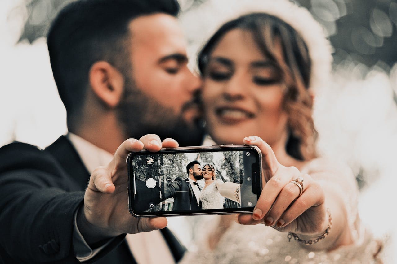 Bride and groom in wedding attire holding a smartphone together to take a selfie outdoors